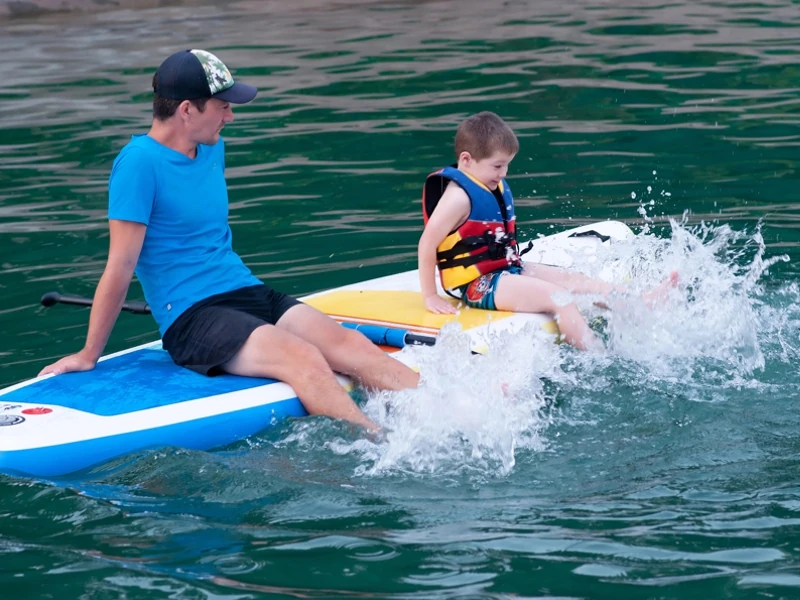 Un père et son fils sont assis sur un paddle, les pieds dans l'eau. Ils agitent leurs jambes pour faire des remous