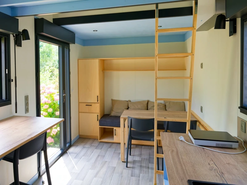 Vue intérieure d'une tiny house : un espace détente avec banquette et coussins, table et chaise ainsi que des rangements. Les fenêtres et baie vitrée offrent une belle luminosité à l'intérieur